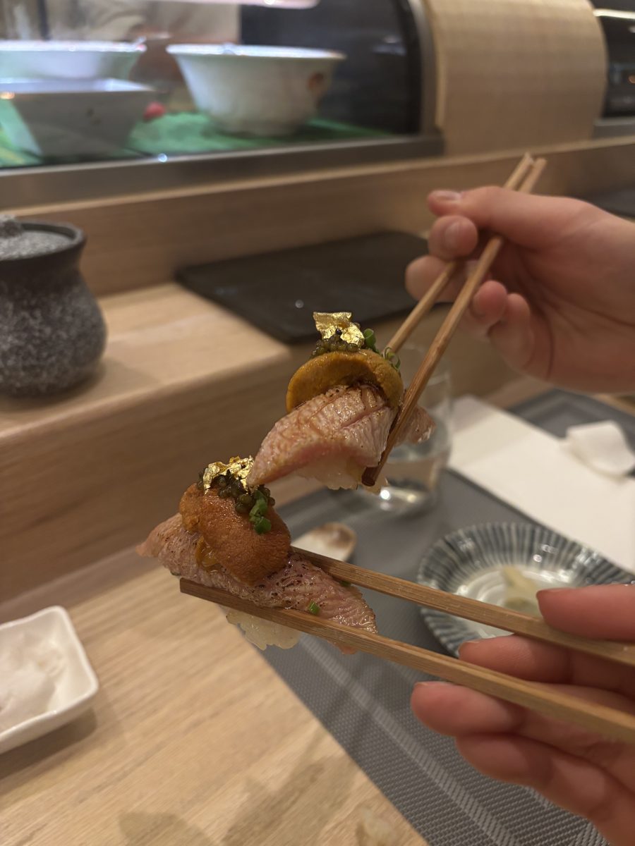 Chopsticks holding a seared nigiri topped with gold leaf at a sushi counter